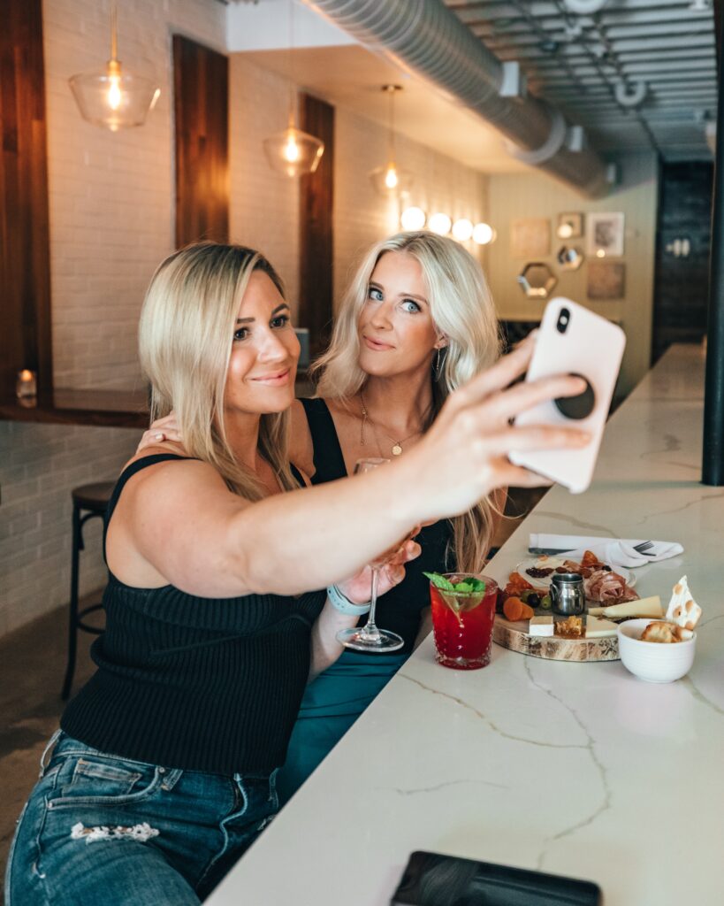 2 women on a night out, taking a selfie. They are having fun, look good all dressed up. Having some food and drink.