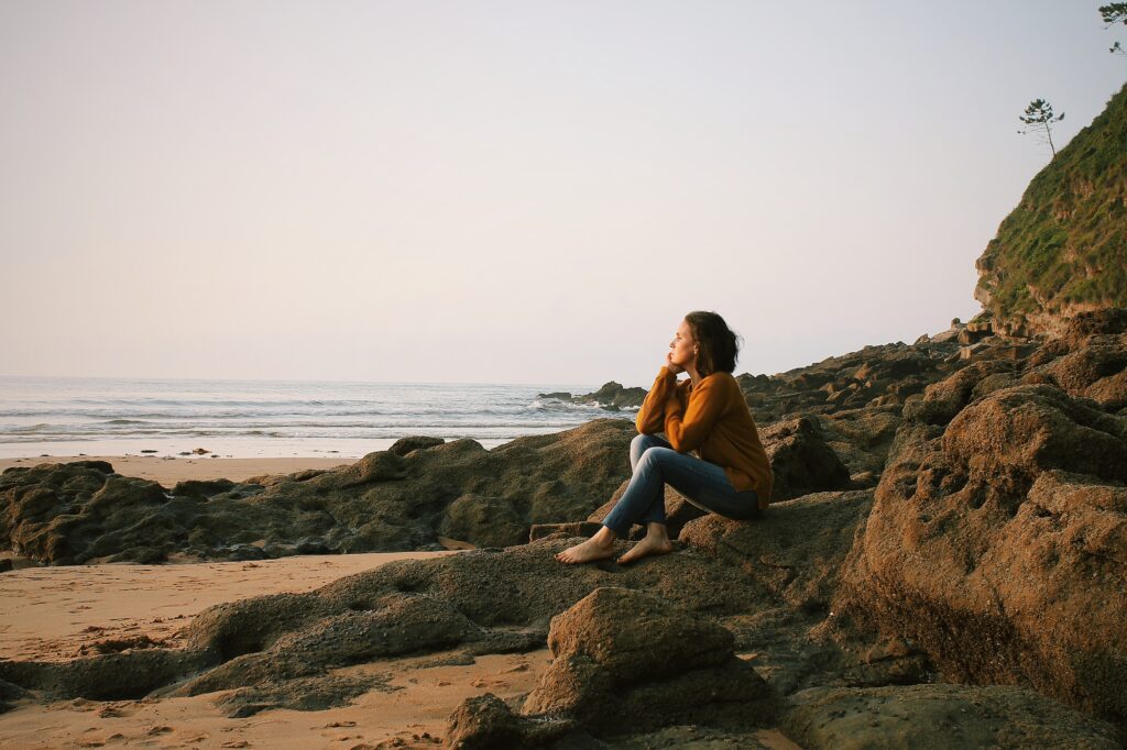 Woman sitting on rocks by the beach thinking if her thoughts are sabotaging her. Photo by Rebe Pascual on Unsplash