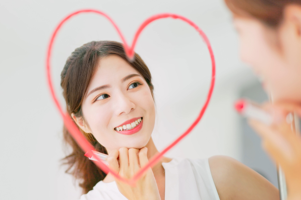 Woman looking in mirror and having drawn a red heart where her face is. She is smiling and appreciating herself. 